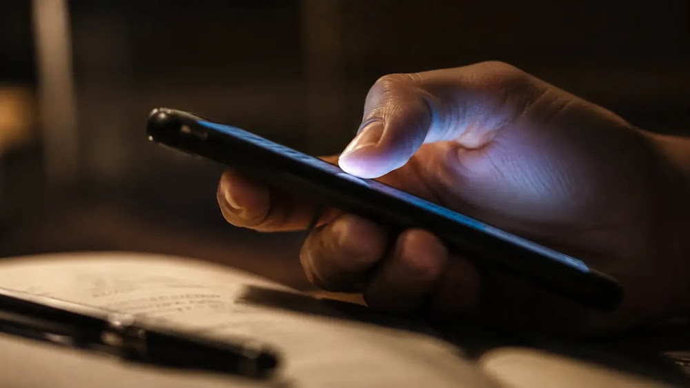 Dispositivos y comportamiento: pulgar en gesto de scroll sobre móvil, libro y bolígrafo abandonados desenfocados al borde inferior